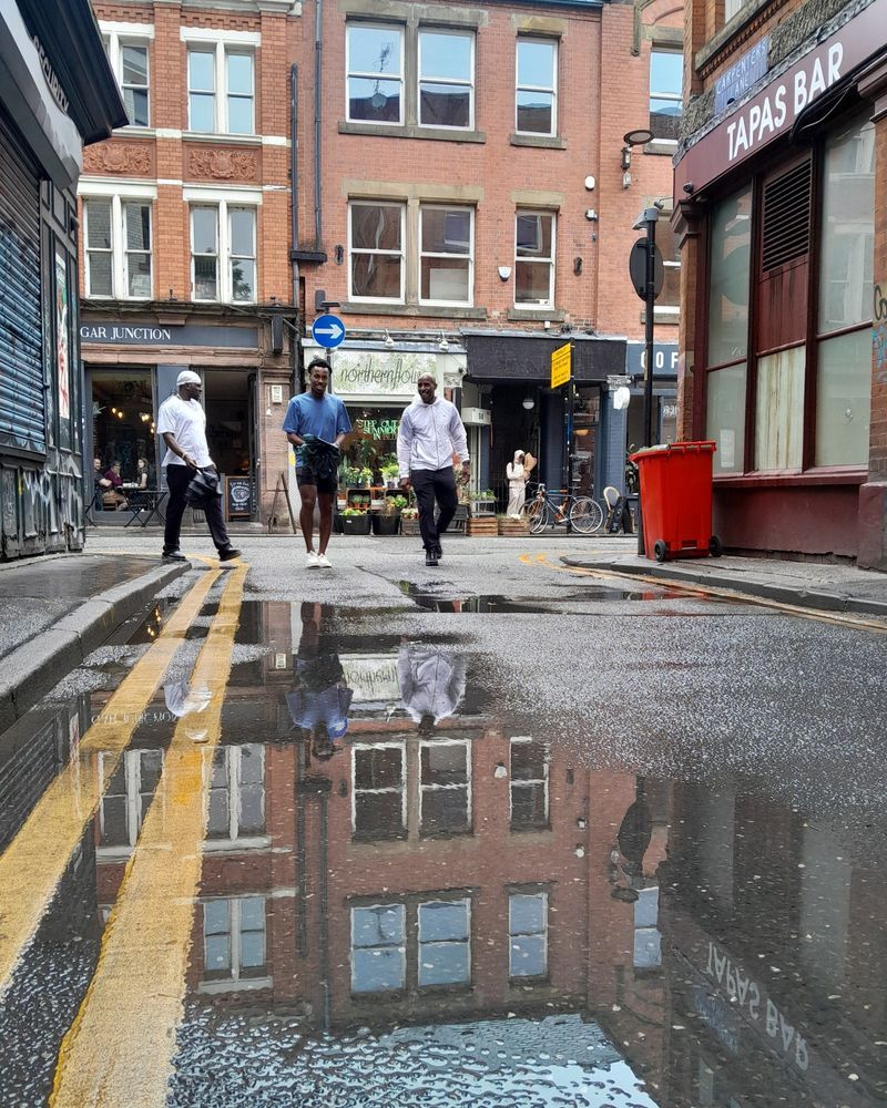 The view from a side alley in Manchester after the rain. A large puddle in the foreground. A florist shop in the distance with a café next door. Someone is stood outside holding a bunch of flowers.  People are gazing out of the cafe window. Three young men are walking down the alley as I take the photo laughing and chatting to each other.