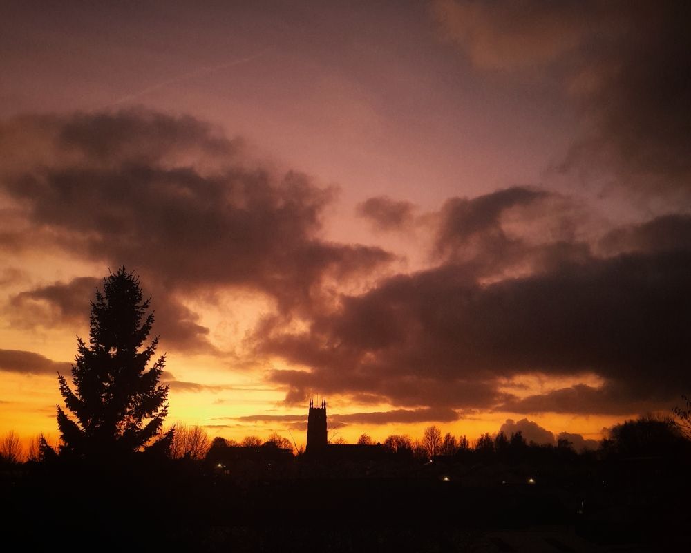 A view from the glamorous local tram station carpark last November. 
On the left a tall fir tree is a silhouette in the dark. In the distance is St Thomas's church surrounded by trees also silhouetted in the darkness. The golden sun was setting with grey clouds above in a sky of orange and pink 