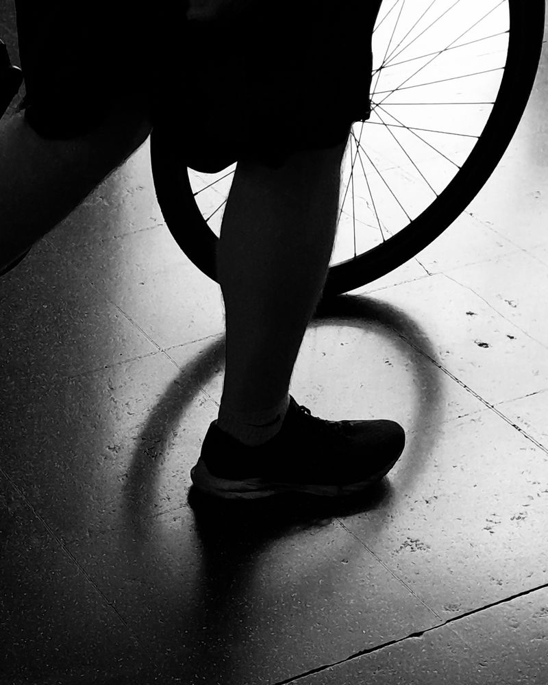 A black and white photo of someone walking past pushing a bicycle. Only one leg and the front wheel is in shot. They are silhouettes with the light from a window behind.  The shadow of the bicycle wheel falling onto the concrete floor 