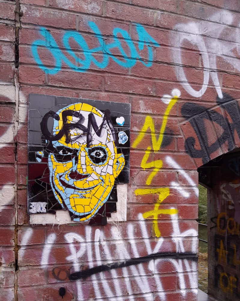 A brick wall that is part of a small bridge over the canal at Castlefield.  It is covered in random graffiti.  On the left is a mosaic of the head of Uncle Fester from the Adam's family. It is a bald man smiling with large staring eyes. The mosaic is mostly bright yellow with a few pale blue and white pieces.