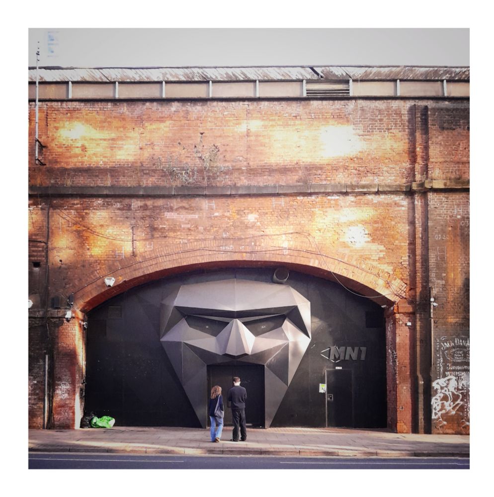 Looking across a street in Manchester where an entrance to a club is under a railway arch. It is a black 3D face of a cyborg and two people are stood looking at the door 