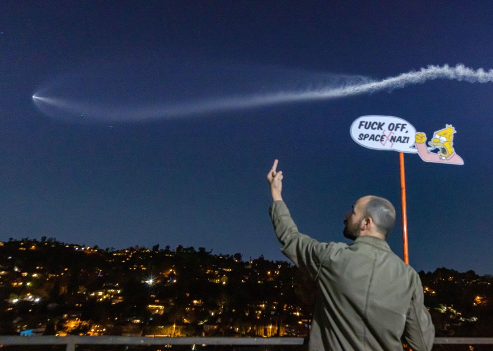 Photo of me holding a cutout protest sign of the “old man yells at cloud” Simpsons meme with a bubble that reads “Fuck off space Nazi”. I’m giving the middle finger to a bright bubble of exhaust in the night sky from a rocket launch that looks like an enormous whale or a celestial sperm.