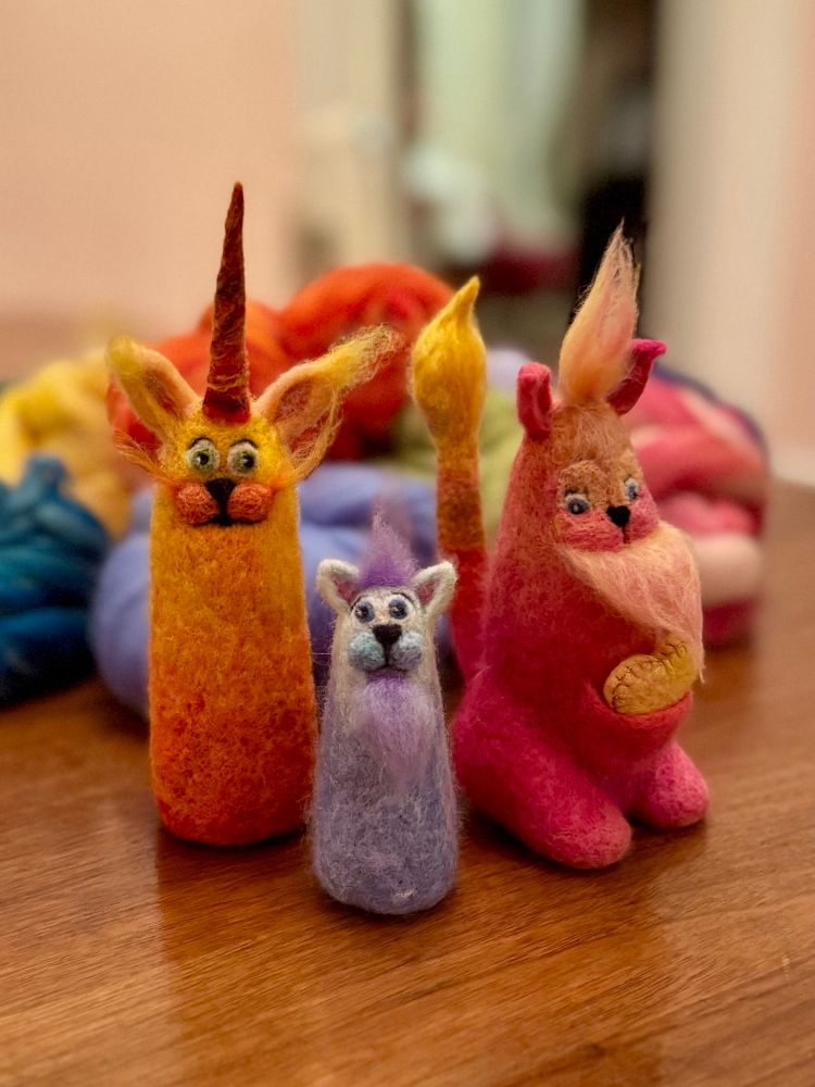 Three needle felted creatures: the pink and yellow one from the previous photo, with striped and tufted tail visible, along with a smaller blue and purple creature and a similar height yellow and orange creature with a long horn.