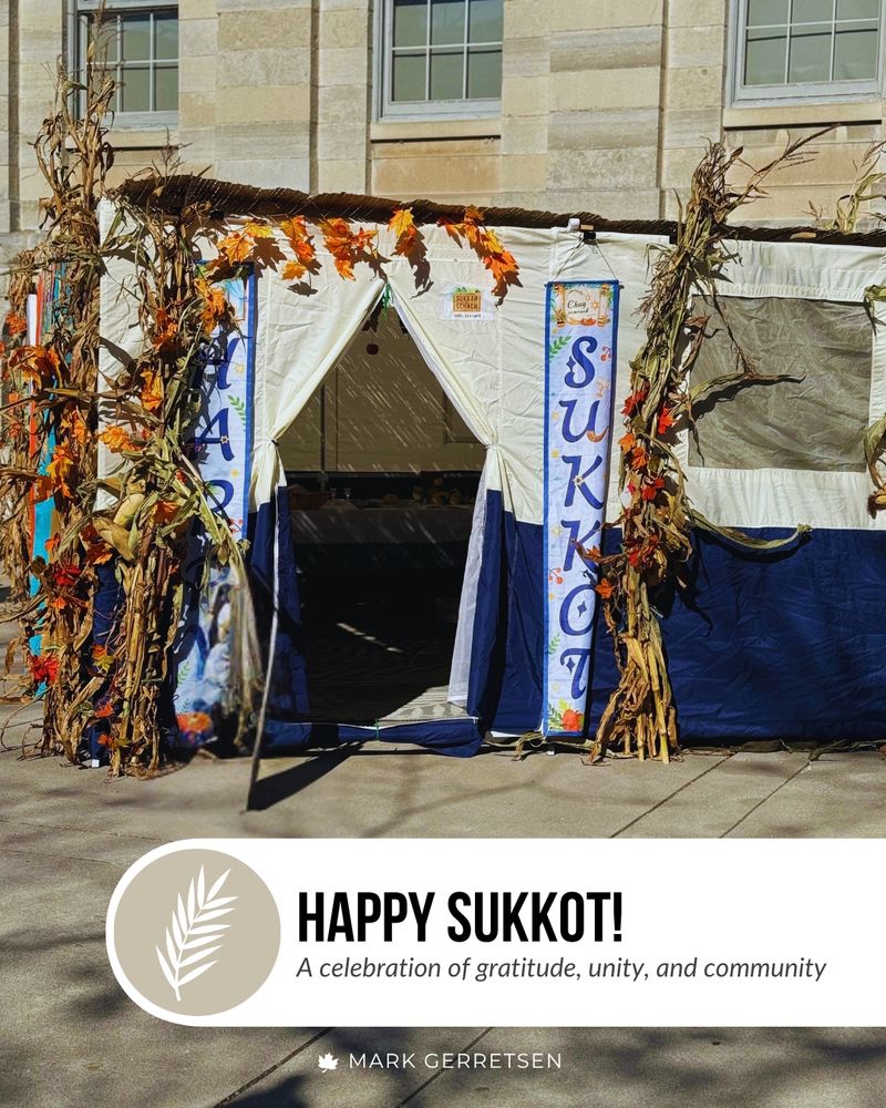 A decorated sukkah structure outdoors, made of fabric panels and adorned with corn stalks and autumn leaves. Two vertical banners on the entrance read ‘Sukkot.’ Below the image, a banner says: ‘Happy Sukkot! A celebration of gratitude, unity, and community,’ with a white palm branch icon and the name Mark Gerretsen.