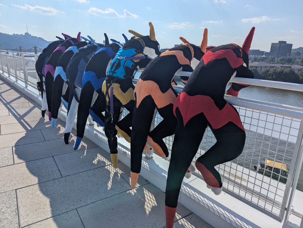 A row of human sized dragon plush from left to right of a water themed followed by a purple, cyan, water themed, dark blue, yellow, orange and red