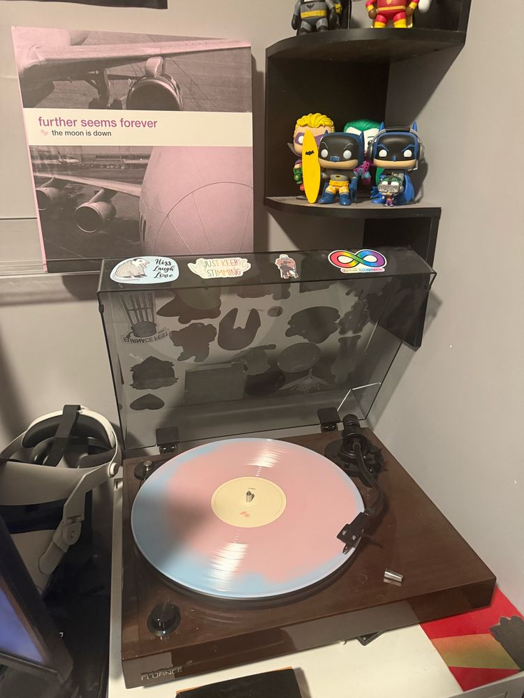 A wood grained record player has its dust cover open. A light blue vinyl record with pink as a secondary color is being played on the record player. Above it is the album art for the record, a pink photograph of a passenger airplane titled “The Moon is Down” by Further Seems Forever.