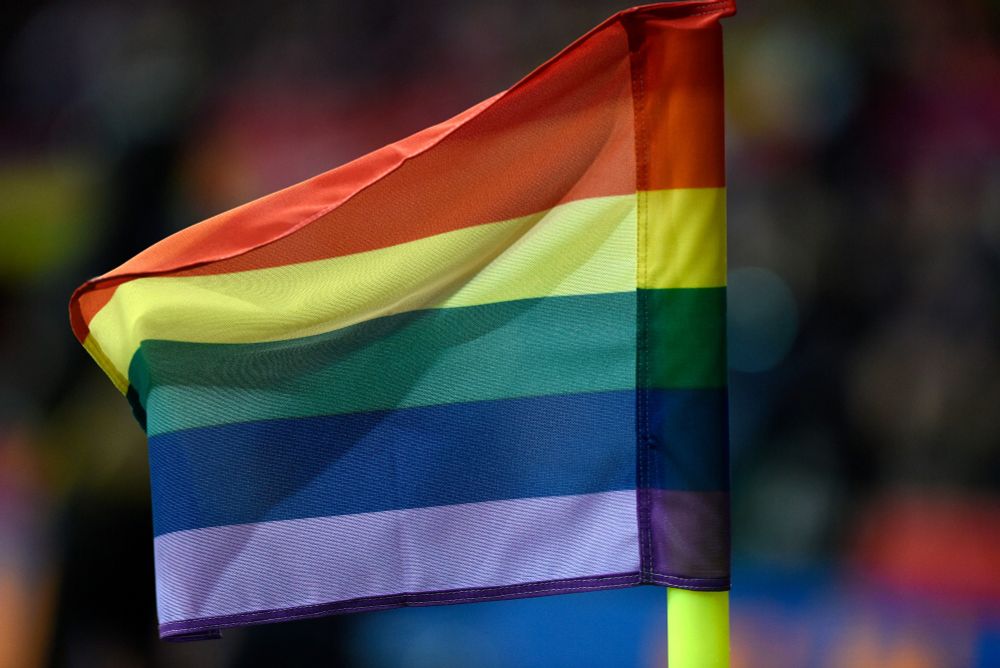 Auf dem Bild ist eine Regenbogenflagge zentral zu sehen, die auf einem neongelben Pfosten gehisst ist. Der Hintergrund ist verschwommen und nicht sichtbar.