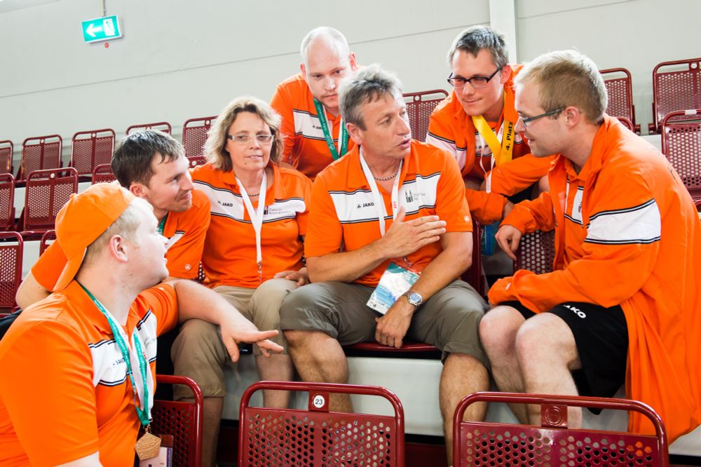 Eine Gruppe von Menschen sitzt auf der Tribüne einer Turnhalle auf roten Sitzen. Es sind sieben Personen, die zusammen sitzen und einer mittig positionierten Person zuhören. Sie alle tragen orangene Vereinsjacken oder T-Shirts. 