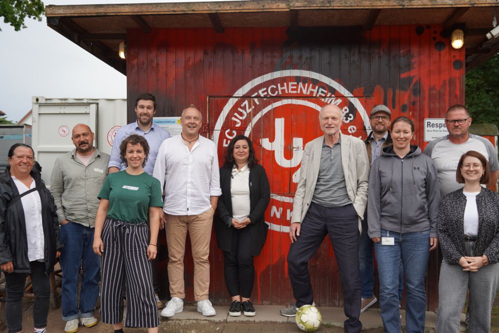 Die Besucher*innen des BAMF und des DOSB stehen zusammen mit den Vertreter*innen des Vereins FC JUZ Fechenheim vor dem Vereinslogo.