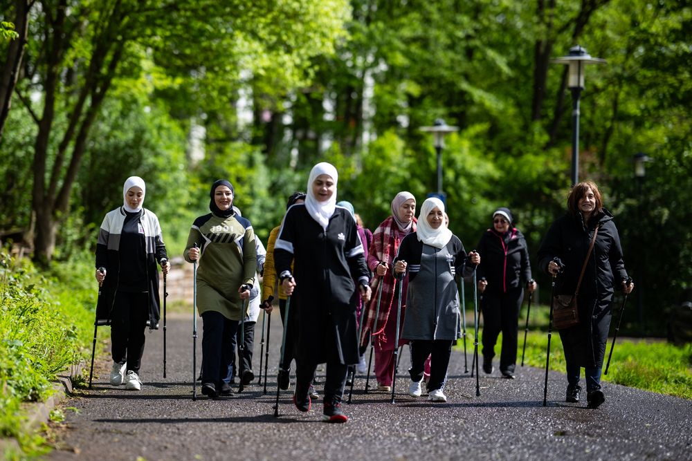 Eine Gruppe von acht Frauen mit Hijab laufen mit Walking-Stöcken in den Händen auf einer Straße in einem grün bewaldeten Park. 