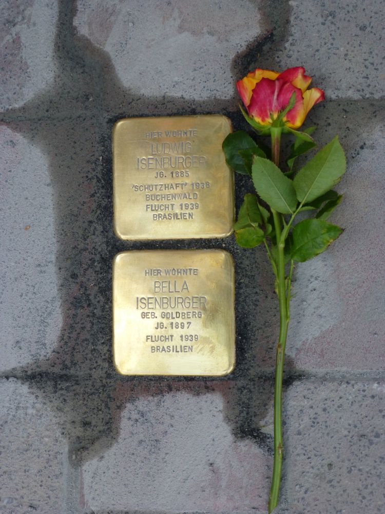Zwei vertikal angeordnete Stolpersteine sind zu sehen im Asphalt. Der obere Stolperstein ist Ludwig Isenburger gewidmet, der untere Bella Isenburger. Rechts neben den Stolpersteinen ist eine Rose abgelegt. 