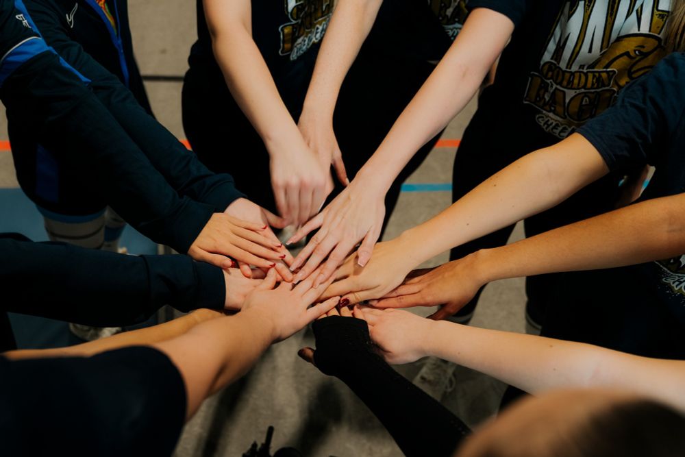 Man sieht die Menschen von verschiedenen Menschen im Mittelpunkt. Die Menschen selbst stehen außerhalb des Rahmens und strecken alles ihre Hände in die Mitte, die sich mit den Handinnenflächen nach unten berühren bzw. aufeinanderliegen.
