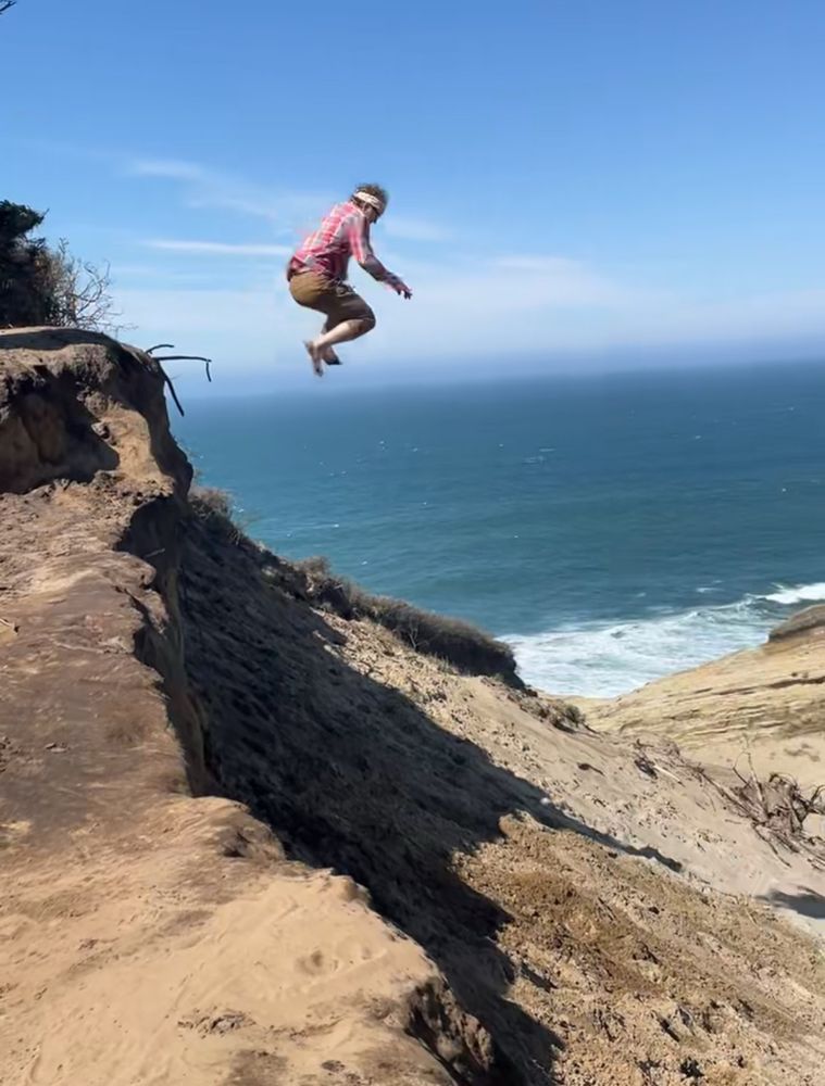 Man in midair jumping off a small cliff into a sand dune with ocean in the backgrounf. 