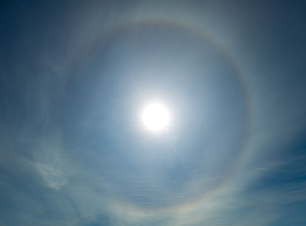 Photograph of the sun surrounded by a bright circular halo, formed by the refraction of light off ice crystals in the atmosphere, against a blue sky with light clouds. Taken in Rancagua, Chile.