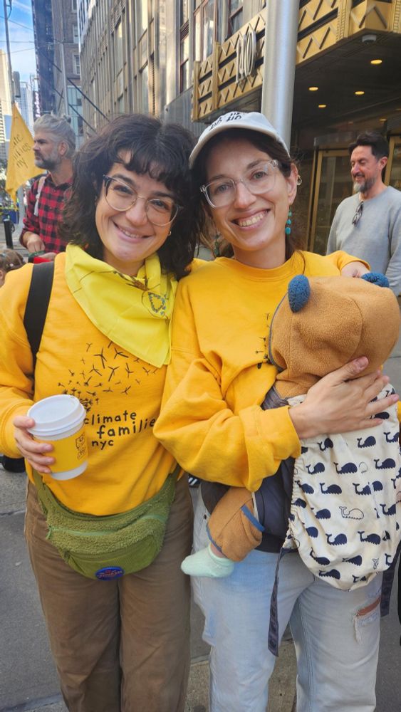 Two women standing together at a protest in yellow sweatshirts one is holding a baby, they are smiling