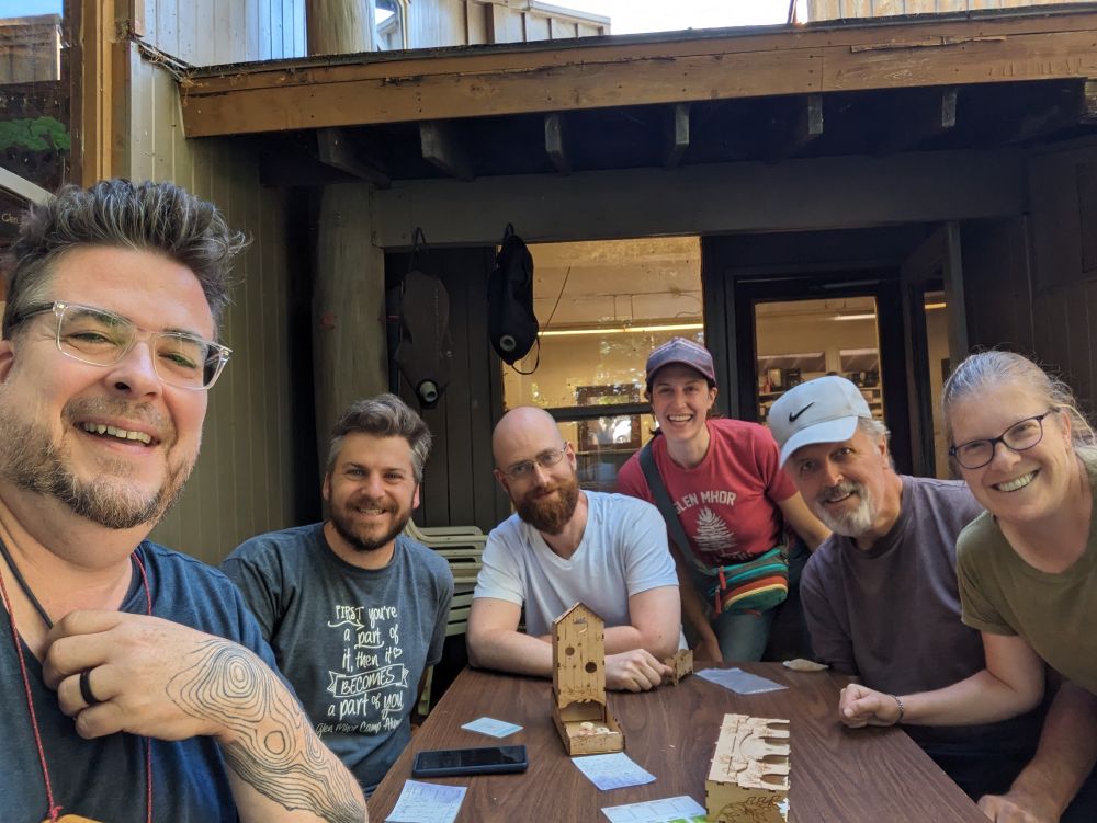 Six board game players posing at the end of a game of Wingspan. They are all outside, gathered around an outdoor table with a summer camp kitchen seen through the windows in the background. 