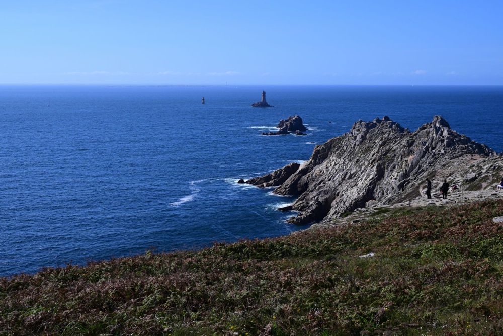 Dunkelblauer Ozean vor hellblauem Himme. Im Vordergrund Felsen, kleine Inselchen mit Leuchtturm.