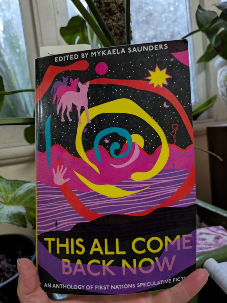 Book being held, backdrop of pot plant in front of window with rain outside. The book cover is colourful shades of pinks, purples, yellow and black. It reads 'Edited by Mykaela Saunders' at the top, with title 'This All Come Back Now' at the bottom and 'An Anthology of First Nations Speculative Fiction underneath