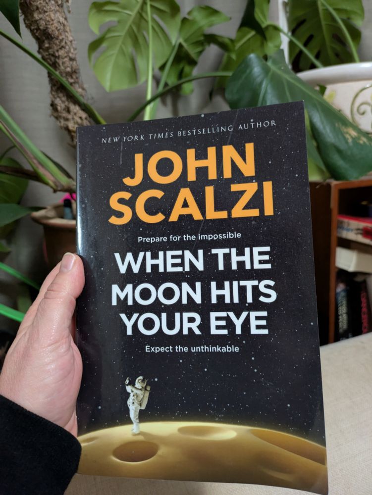 Hand holding a book with pot plants in the background. The author name is in orange all caps san serif font at the top JOHN SCALZI. The title is in white text, again all caps san serif font WHEN THE MOON HITS YOUR EYE. The top three quarters of the background is dark night sky with star twinkles, and at the bottom is an astronaut standing on a very cheese looking surface of the moon