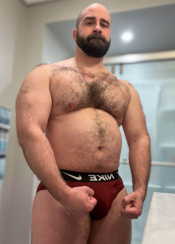 A photo of me in my underwear. A large man  with a hairy chest, arms and shoulders with a bald head and dark beard. Arms are down and flexing slightly. Wearing maroon Nike underwear with a black waistband. Taken in a brightly lit bathroom with a shallow depth of field. 
