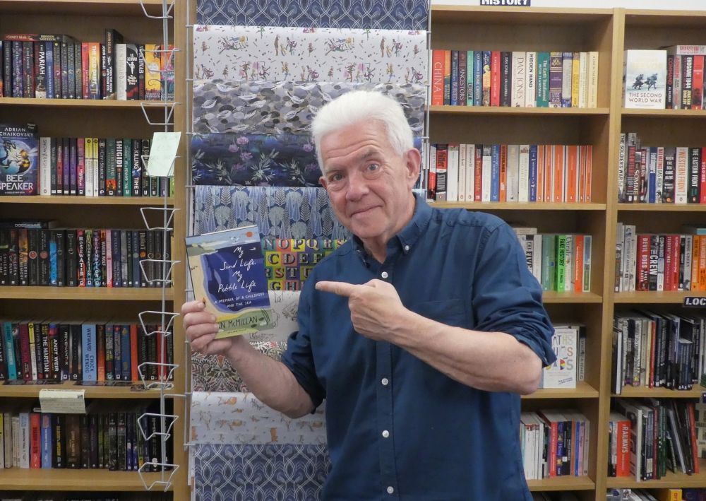 Photo of Ian McMillan, holding and pointing at one of the copies of My Sand Life, My Pebble Life which he had just been signing. A rack of wrapping paper immediately behind Ian.