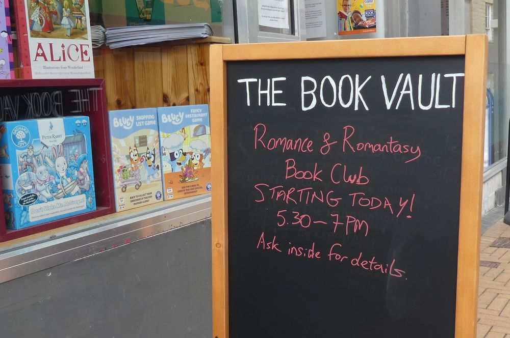 Photo of pavement chalkboard with The Book Vault in white capital letters, then in red Romance and Romantasy Book Club, starting today! 5.30 - 7pm, ask inside for details. Part of the shop window visible to the side with Bluey and Peter Rabbit Orchard Toy games/puzzles, and part of an art book of Alice in Wonderland.