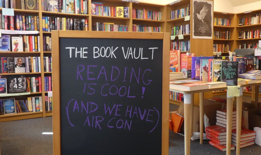 Photo of a pavement chalkboard, with writing - The Book Vault. Reading is Cool! (And We Have Air Con). Taking in store with bookshelves and tables in the background, too much sunlight to take a photo without sign reflection outside!