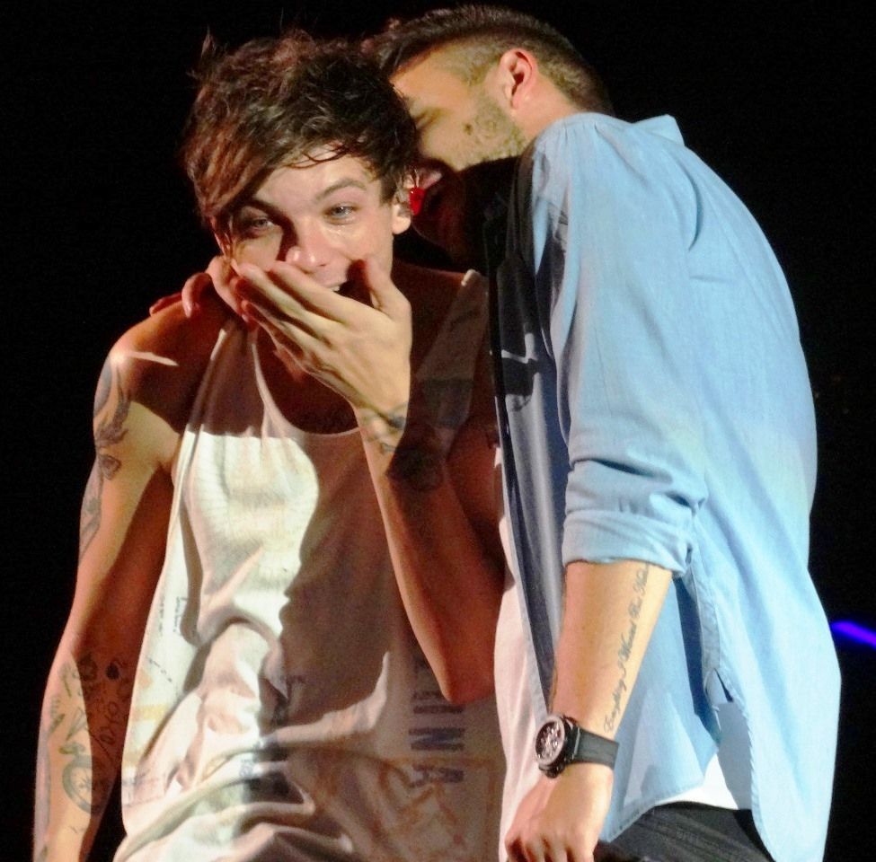 During a One Direction concert, Liam Payne whispers in Louis Tomlinson's ear. Louis has one hand over his mouth as though hearing something delightfully shocking.