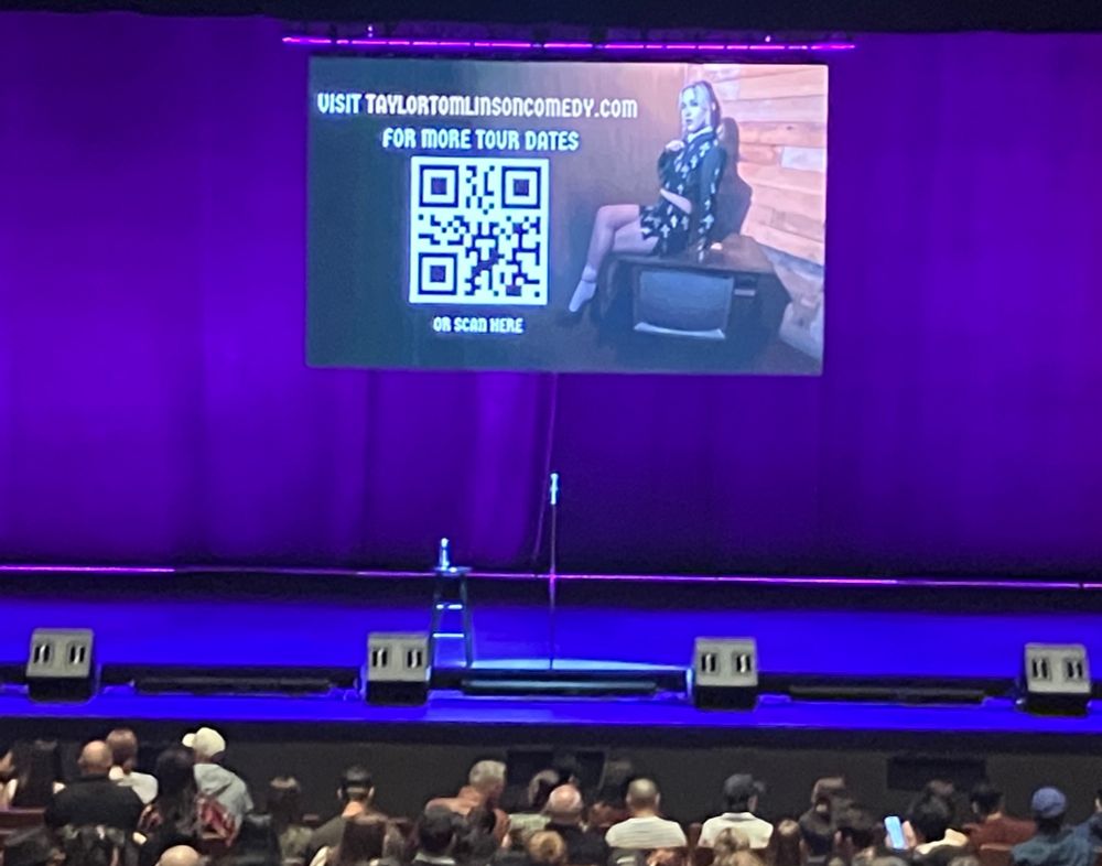 A photo of the stage of the Queen Elizabeth Theatre in Vancouver before the show. We see an image of Taylor Tomlinson on a screen with a qr code for future dates. There is a mic and stool with bottle of water on stage. The curtains have a purple light hue. Front rows are packed with people.