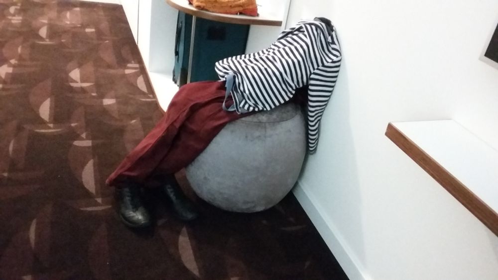 Kleidung, die wie ein erschöpftes Wesen auf einem Sitzhocker lieht, Kragen des Pullovers liegt gegen eine Wand, wie kopflos.
Kleidung  ist gestreifter Pullover, rote Hose, schwarze Schuhe. Hocker grau, Fußboden braun, grafisches Muster