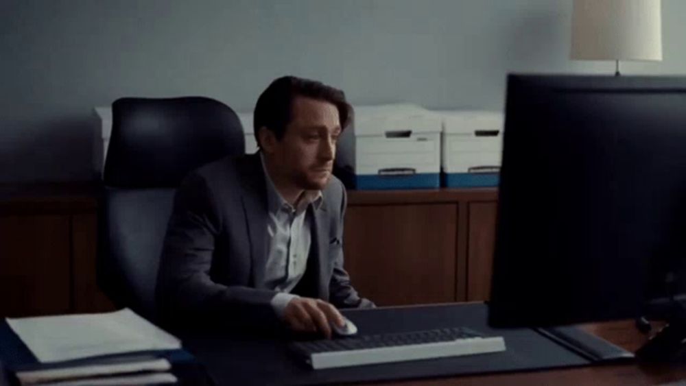 a man in a suit sits at a desk in front of a computer monitor