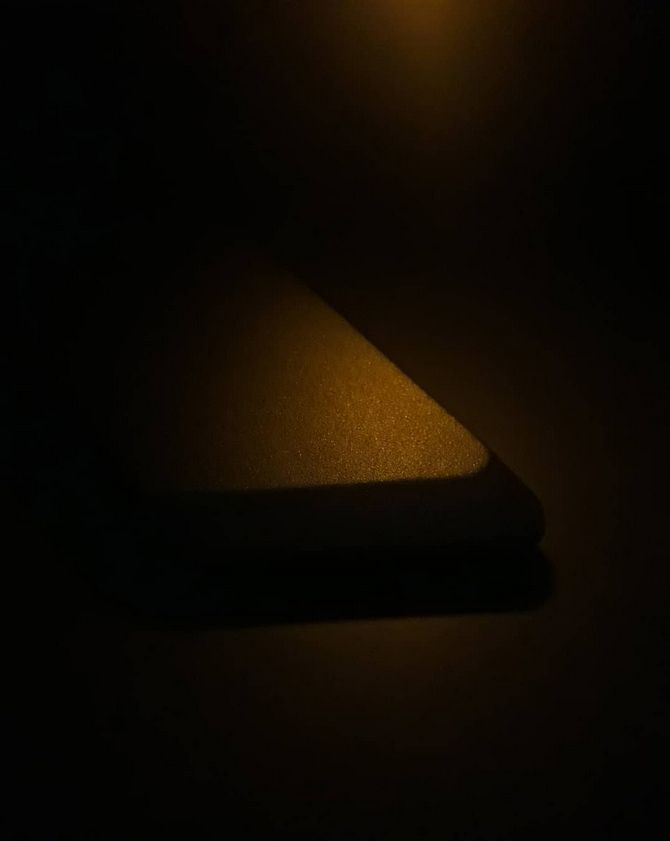 A dimly lit close-up of a rectangular object, possibly a smartphone or a power bank, placed on a surface. The warm, golden light highlights its textured surface and casts a soft shadow, creating a moody and minimalistic composition with a dark background.