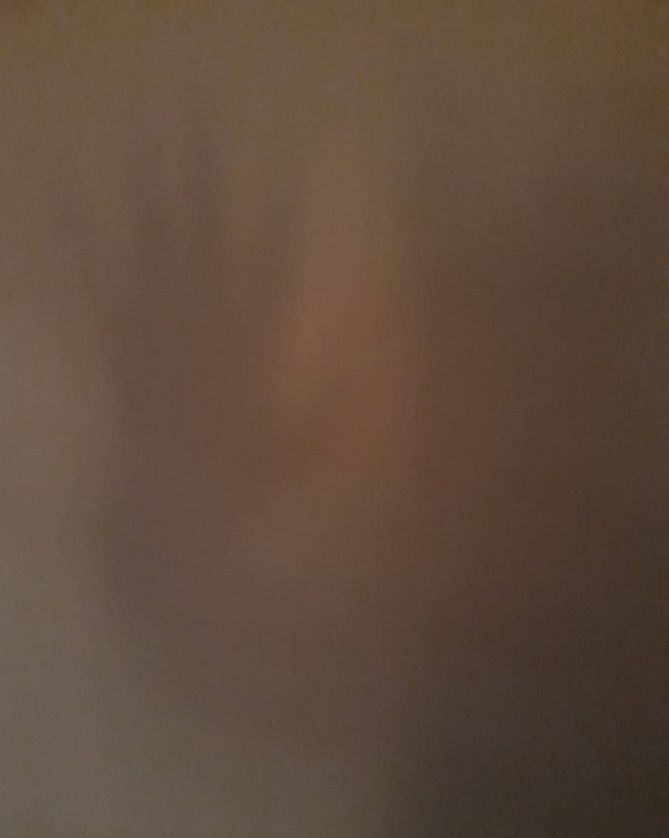 A blurry, dark-toned image with a faint outline of a handprint visible on a surface. The image has a muted brownish color and lacks sharp details.