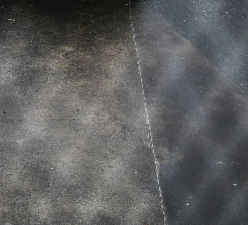 The image shows a wet concrete surface with dark and light patches. There is a visible dividing line between two sections, possibly indicating different materials or levels of wetness. Small white specks and dirt are scattered across the surface. The lighting creates subtle reflections on the wet areas.