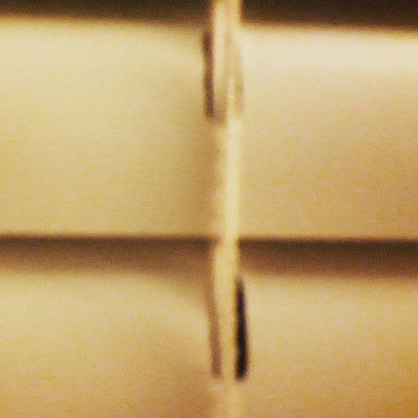 The image appears to be a close-up of window blinds with a vertical cord running through the center. The focus is slightly off, making the details blurry, and the lighting gives the scene a warm, yellowish tone. The cord has dark markings, possibly stitching or wear.