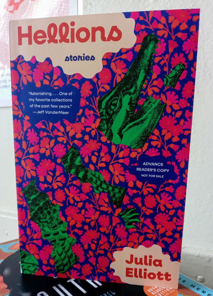 Galley of Hellions standing up on a stack of books. Cover features a drawing if an open mouthed alligator with an overlay of small pink and orange flowers with deep ocean blue underneath. Lettering in red.