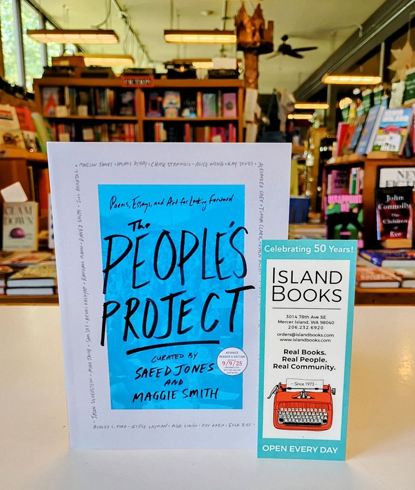 Galley of The People's Project with an Island Books bookmark perched on the front counter at Island Books. The cover is blue with the title and author information in black lettering. Bookmark has a typewriter on it, display tables and shelves are in view.