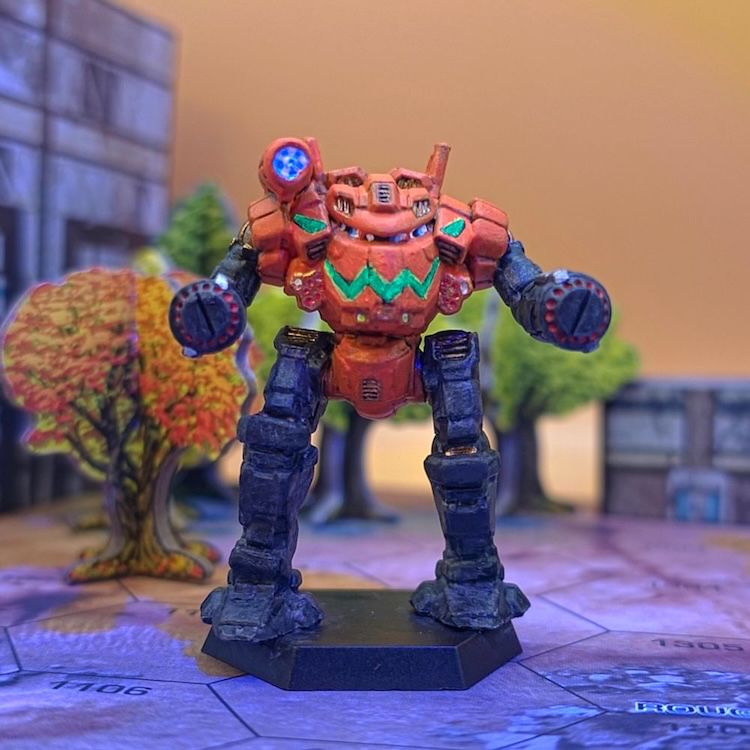 A BattleTech mini - the Shogun - painted like a big jack-o-lantern (the body is orange, the limbs are black, and a fluorescent glowing smile and eyes are painted across the chest) and standing on a hexmap among papercraft trees and buildings.