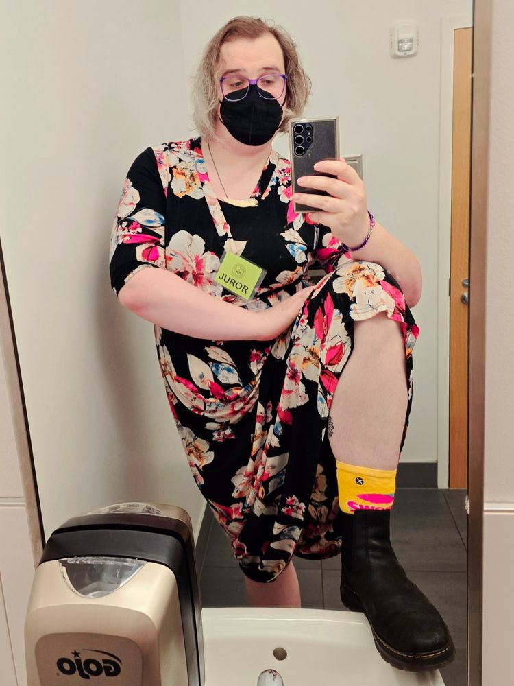 A brunette white woman in purple glasses, a black n95 face mask, and a black maxi dress with a floral print takes a selfie while doing a sink stomp, revealing that her black Doc Martens are accompanied by Swedish Fish socks. She's also wearing a juror's ID badge because there is nowhere better to serve cunt than a courthouse while waiting around during jury duty.
