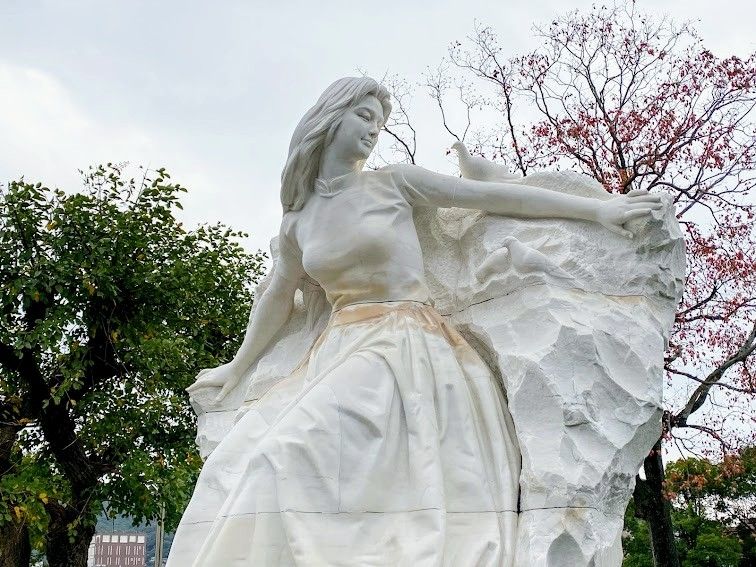 Das Bild zeigt eine weiße Marmorskulptur einer Frau mit langen Haaren, die seitlich sitzt oder sich nach vorne beugt. Sie trägt ein langes, fließendes Kleid, und ihr rechter Arm ist ausgestreckt, auf dem eine Taube sitzt. Der Hintergrund zeigt Bäume, teils mit grünen, teils mit roten Blättern, sowie einen bewölkten Himmel.