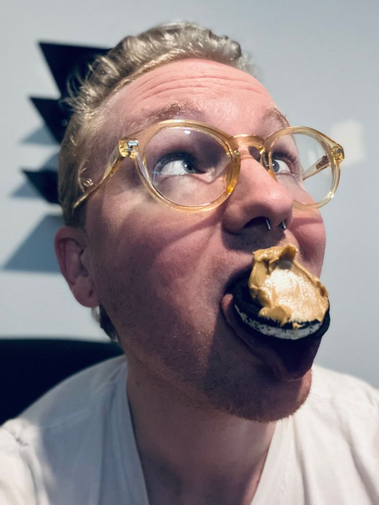 closeup selfie of Fox (white, futch) holding a chocolate sandwich cookie smeared with peanut butter on its tongue, looking upwards and crossing its eyes in ecstasy.