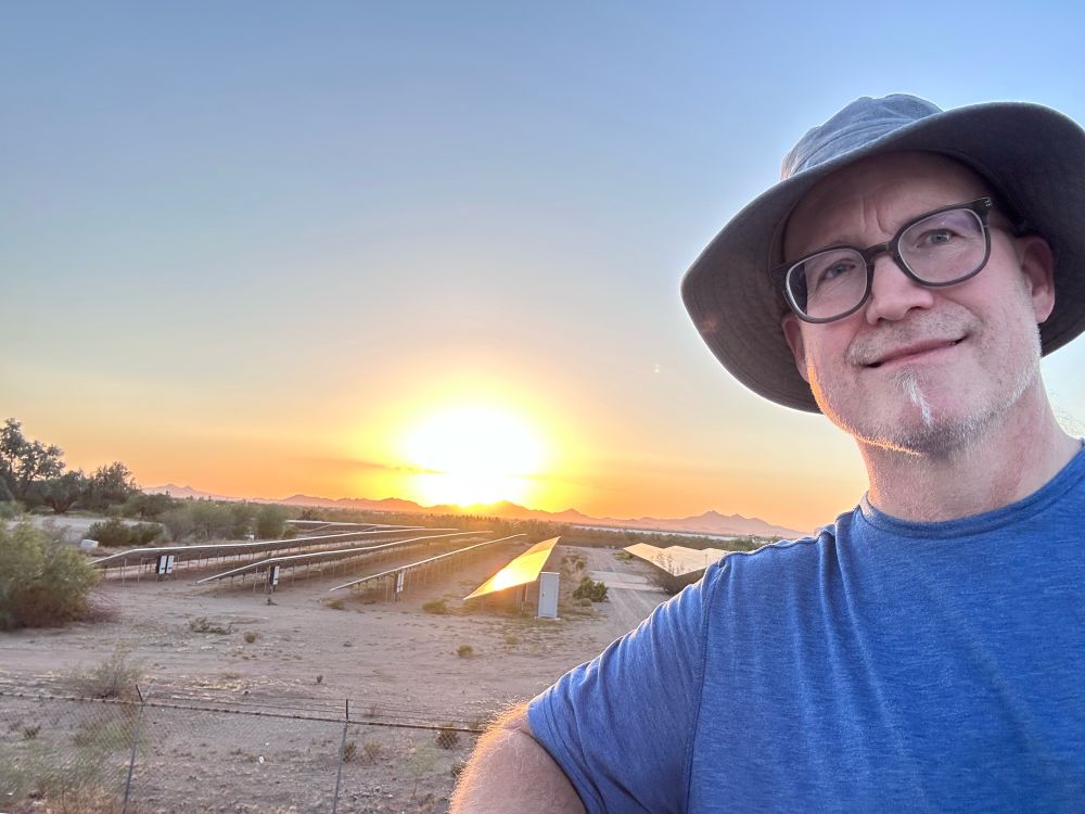 A bespectacled guy wearing a hat posing in front of a solar farm with a beautiful and radiant sunset behind them.