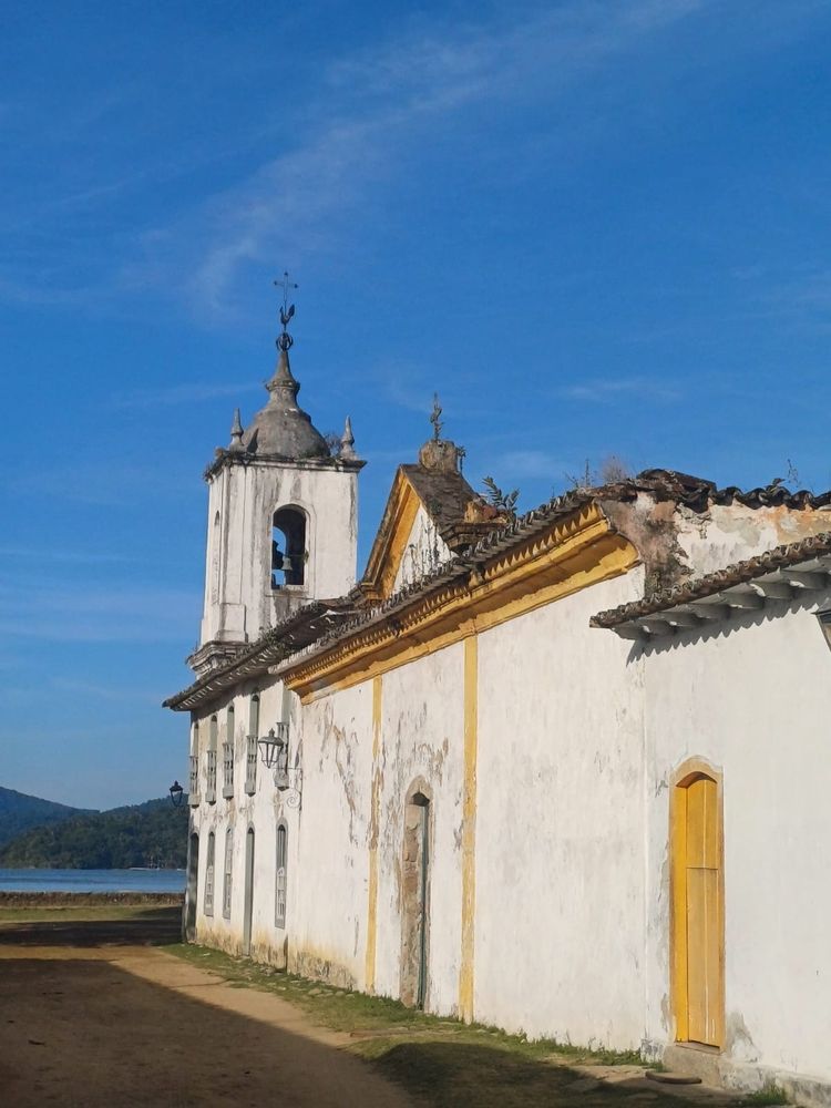 Lateral de uma igreja de frente para o mar em Paraty. Não tomo honesto uma cúpula acinzentada sino suas cores são brancas acinzentado e amarelo bem forte a passagem é de terra e Pedras. Tá de frente ao mar azul e a dois morros menores bem esverdeados.