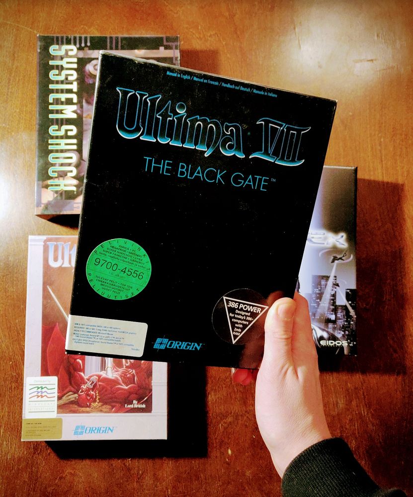 Photo of three PC game boxes on a table (System Shock, Ultima VI, Deus Ex). Hand holding a fourth box (Ultima VII) above the others.