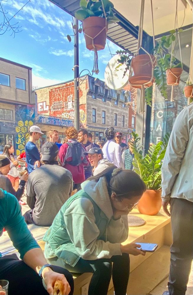 A young woman looks down at her phone while sitting by the window of a busy cafe. She is surrounded by a group of people socializing and engaging with each other in a lively discussion. They are illuminated by the light of a bright sunny day, while she sits in her own shadow, facing away from the window, preferring the glow of her screen. 