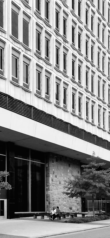 A couple sits on a bench below a building with many concrete windows. Black and White 