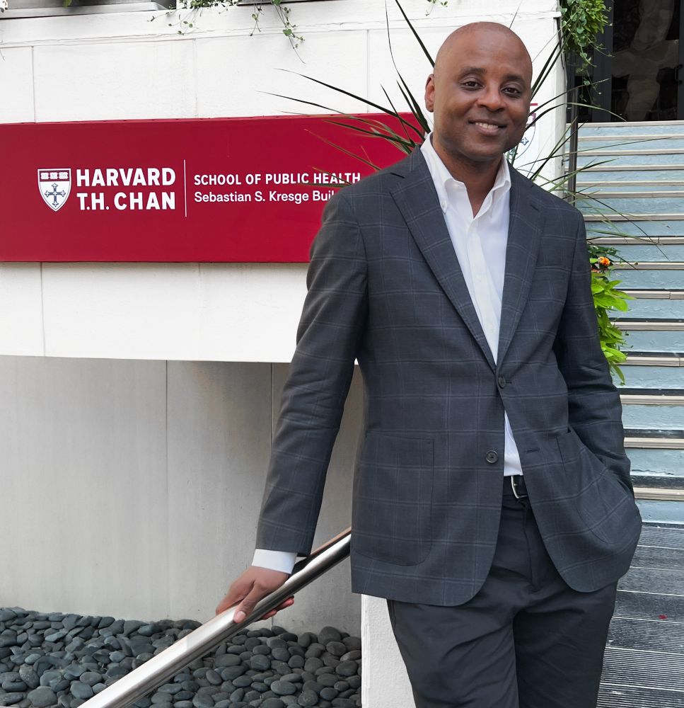Kareem Carr standing outside Harvard School of Public Health moments after his successful PhD defense. He is smiling, dressed in a dark-patterned sport coat and dress pants, leaning casually against a railing. There is a bright red sign over his right shoulder that reads “Harvard T.H. Chan School of Public Health”.