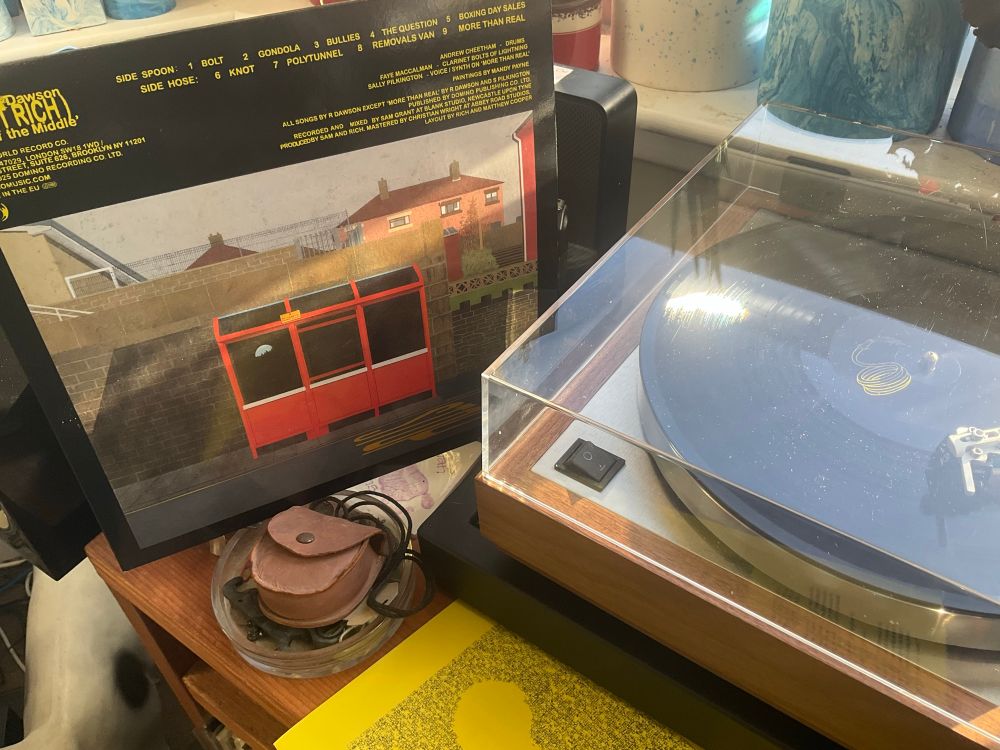 Richard Dawson album cover with a painting by Mandy Payne of a red Bus Stop in front of a stone wall, leaning against a speaker behind it and a turntable to the right of the sleeve. In front of the turntable is the lyric booklet printed on yellow paper.