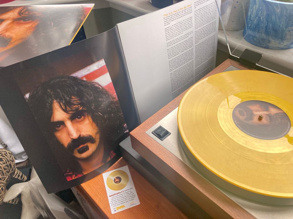 On the a turntable with yellow snow coloured vinyl record showing the a side label with the portrait photo of Frank Zappa from the album cover. On the left lying on a speaker is the album cover and resting against the speaker is the accompanying booklet showing an uncropped version of the cover photo.