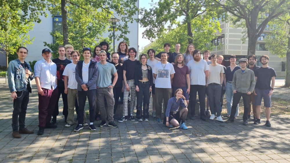 Gruppen Bild des Fachschaftsrat Mathematik Physik. Im Hintergrund Bäume und 2 Häuser. Das linke Grau, das rechte Golden.